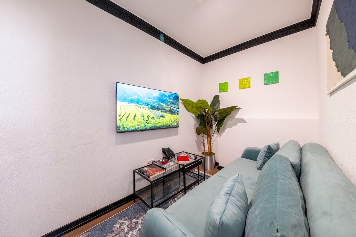 The living area features a comfortable green sofa positioned next to a glass coffee table. A flat-screen television is mounted on the wall across from the seating. Decorative wall art in shades of green adds a touch of color, while a potted plant contributes to the relaxed ambiance.