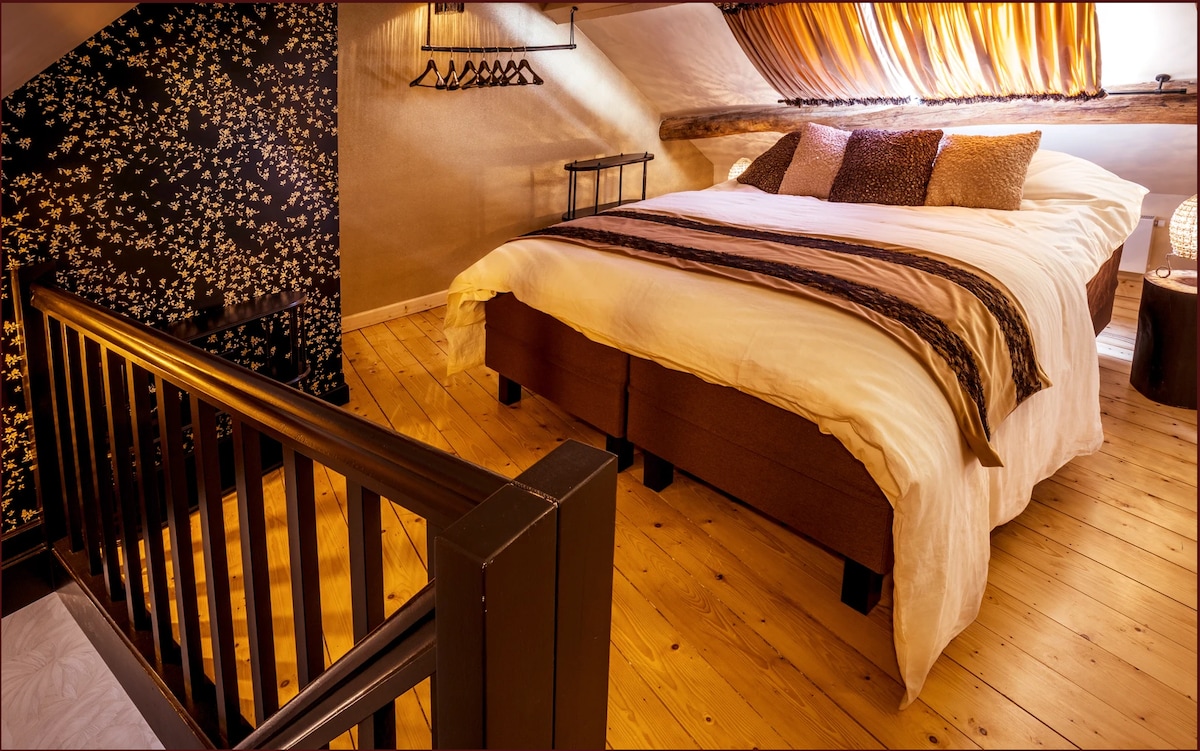 A sleeping area features a spacious bed adorned with decorative pillows. A patterned accent wall complements wooden flooring. Soft lighting creates a warm ambiance, while a wooden railing and hooks for clothing are visible in the background.