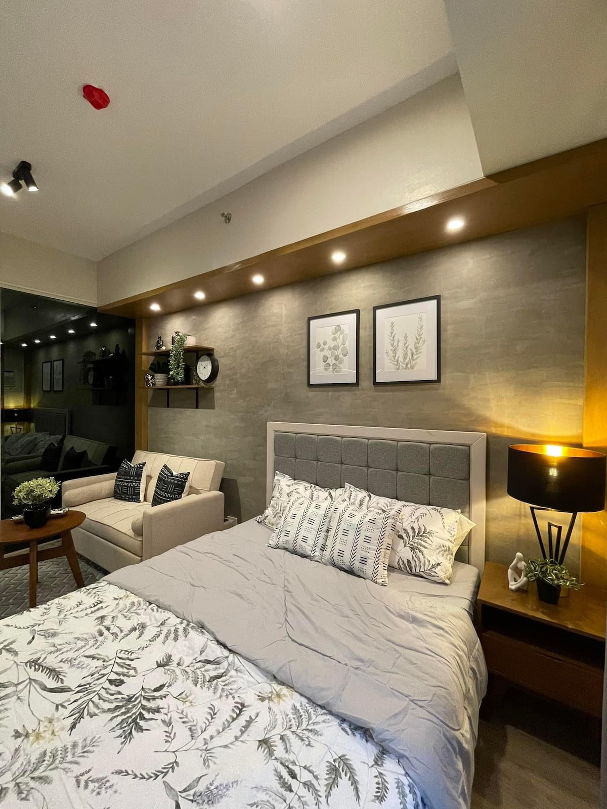A bedroom area features a comfortable queen-sized bed with decorative pillows and a subtle floral-patterned blanket. A cozy two-seater sofa is positioned nearby, accompanied by a side table. Soft lighting highlights the artwork on the wall, creating a peaceful ambiance.