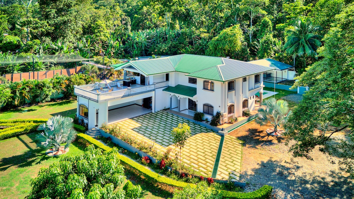 The property is depicted from an aerial view, showcasing a spacious two-story building surrounded by lush greenery. Well-maintained pathways lead to the entrance, with outdoor seating areas visible on the upper balcony. A covered parking area is located to the left, accommodating multiple vehicles.