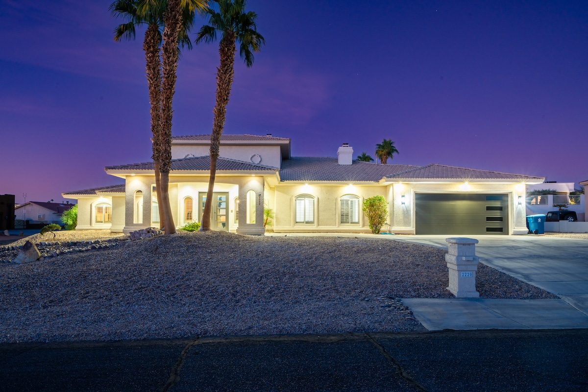 A spacious single-story home is set against a twilight sky, featuring a paved driveway leading to a three-car garage. Lush palm trees frame the entrance, and soft lighting highlights the exterior architecture, creating a welcoming atmosphere.