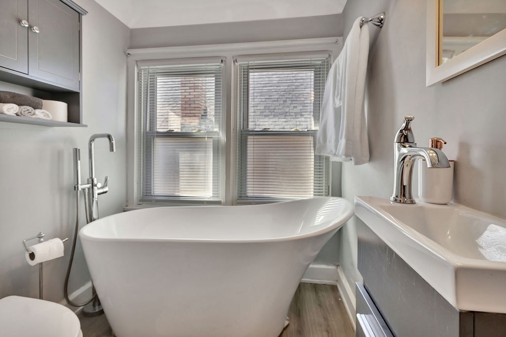 A bright bathroom features a freestanding soaking tub positioned between two windows, allowing natural light to fill the space. A sleek vanity with a modern sink is located beside the tub, with neatly stacked towels stored nearby, contributing to an organized appearance.