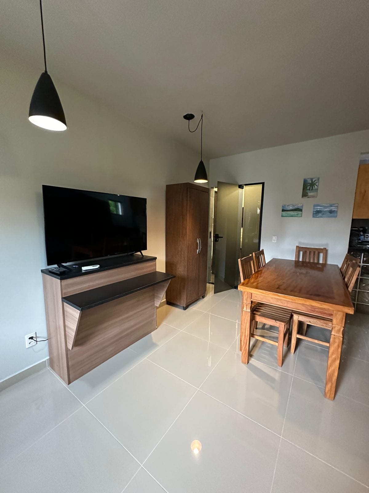 The dining area features a wooden table with six chairs, positioned near a modern television mounted on a low cabinet. The space is enhanced by pendant lighting above, and a door leads to another room. Bright tile flooring reflects natural light from nearby windows.
