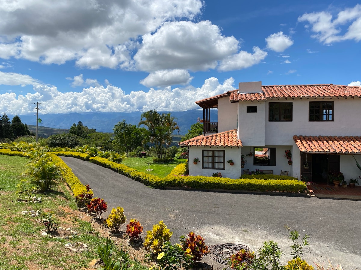 A charming two-story finca is positioned against a backdrop of rolling hills and vibrant skies. The entrance features a winding driveway bordered by colorful plants. Lush greenery surrounds the property, enhancing the serene setting and offering expansive views of the natural landscape.