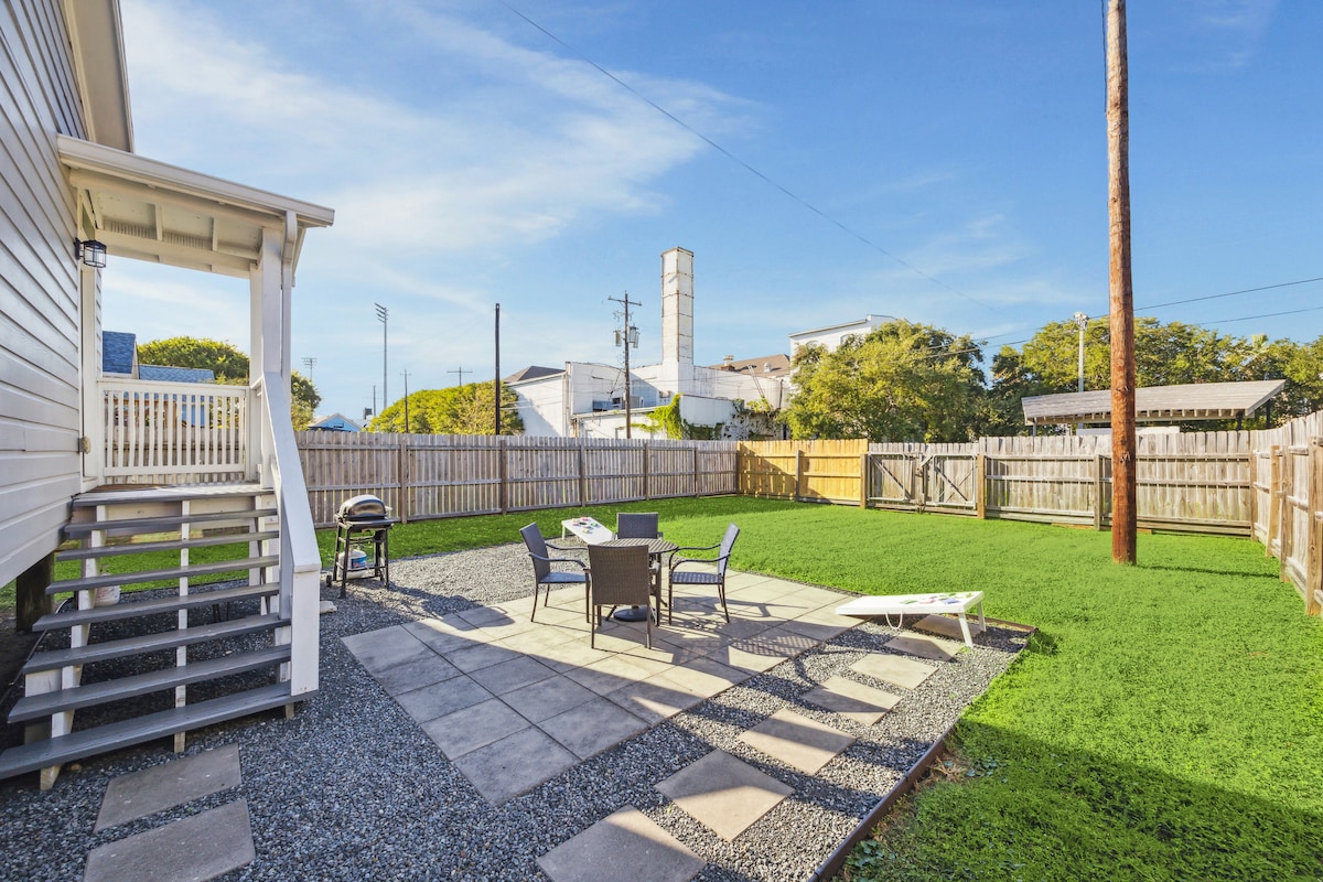 A spacious backyard patio area is shown, featuring a stone-paved surface with outdoor furniture arranged around a table. Lush green grass surrounds the space, complemented by a wooden fence enclosing the yard. The entrance steps lead up to the bungalow, enhancing accessibility.