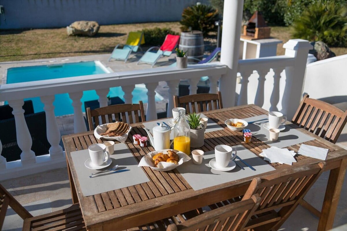 An outdoor dining table is set with cups, plates, and a pitcher, surrounded by wooden chairs. In the background, the inviting pool reflects sunlight, accompanied by loungers and a barbecue area. The scene is enhanced by greenery, creating a calm atmosphere for meals.