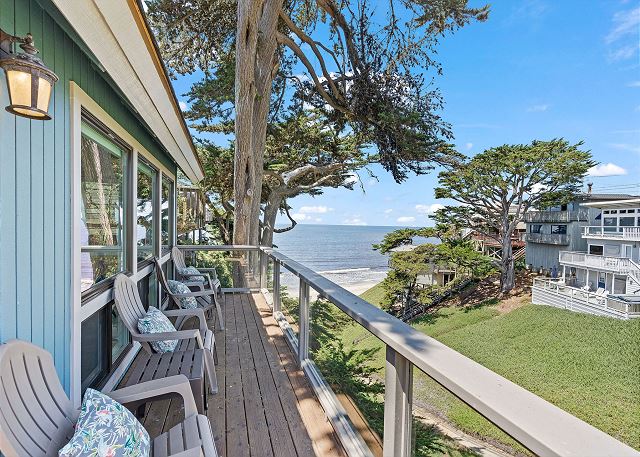 A spacious deck is shown with several comfortable chairs arranged for ocean viewing. Lush greenery and a gentle slope lead to the beach. The ocean sparkles under a clear blue sky, with trees providing natural shade around the seating area.