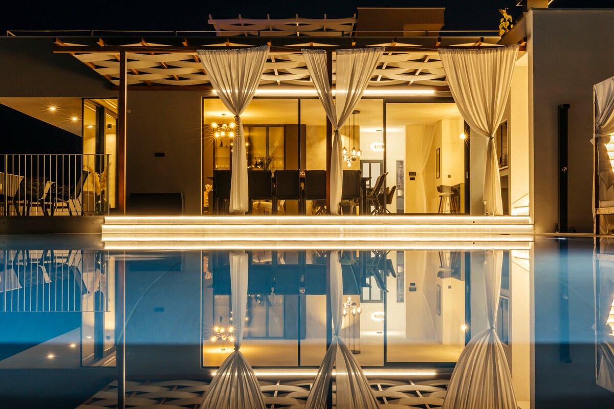 The villa exterior is illuminated at night, showcasing elegant draped curtains over large glass doors. The reflection of the structure is visible in the calm waters of the pool, creating a serene and balanced visual effect.