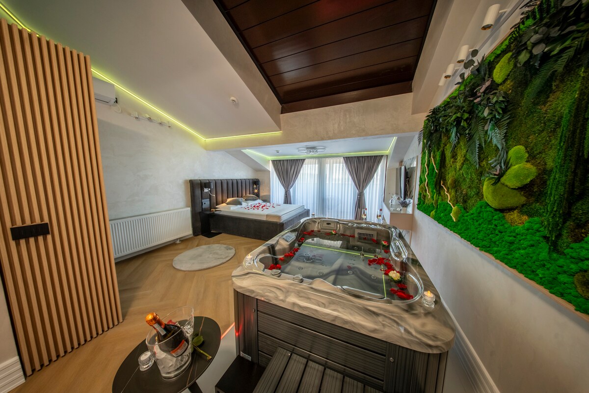 A spacious room features a luxurious hot tub adorned with rose petals. A textured green wall adds a natural touch beside a well-appointed sleeping area with ambient lighting. Large windows provide natural light, complemented by soft drapes hanging elegantly.