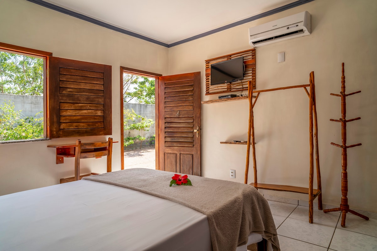 A neatly arranged room features a spacious bed with a neutral-colored blanket. Natural light enters through two windows, framed with wooden shutters. A mounted television is positioned on the wall, complemented by a rustic wooden clothes rack and a small seating area near the entrance.