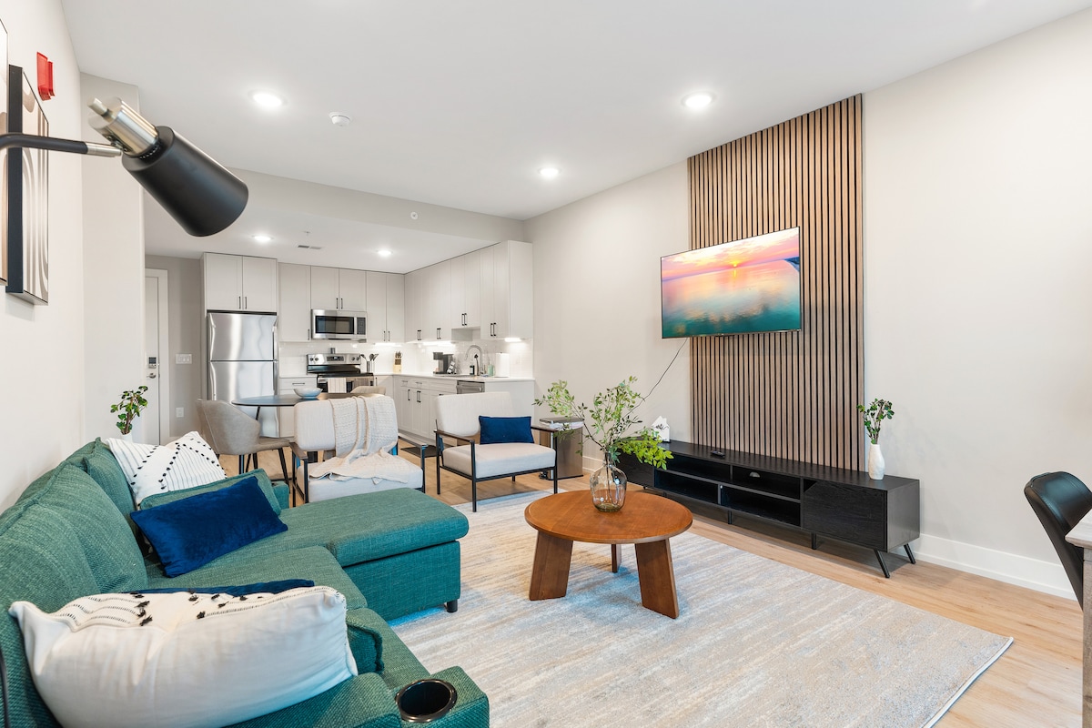 An open concept living area features a sectional sofa in a teal hue, complemented by a round wooden coffee table. A 50-inch Roku Smart TV is mounted on the wall, while a well-equipped kitchen with modern appliances is visible in the background.