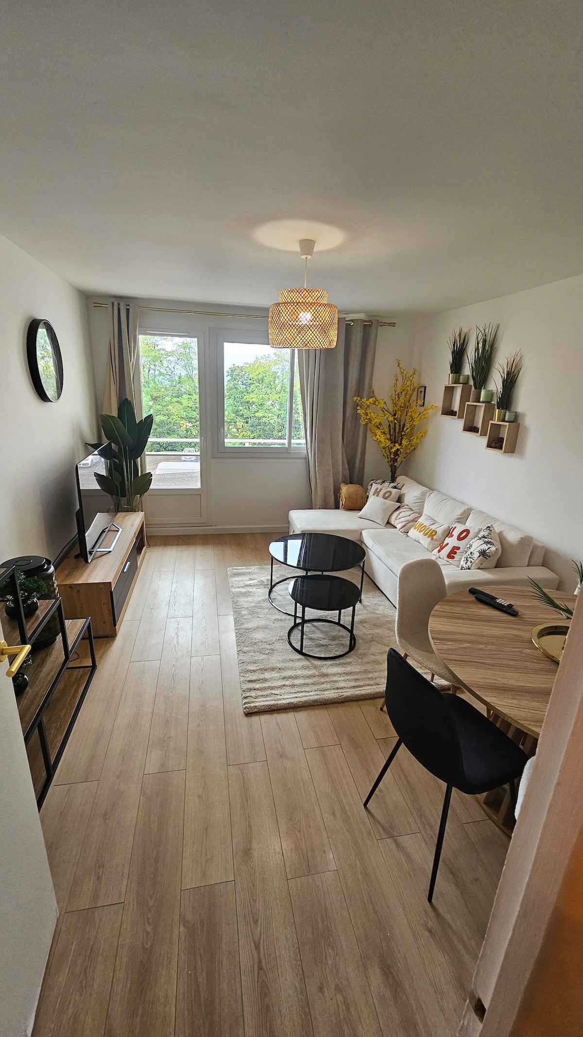 A bright living area is featured, showcasing a light-colored sofa adorned with decorative pillows. A round black table is positioned in front, surrounded by stylish chairs. Natural light streams through large windows, complemented by decorative plants and wall-mounted shelves with greenery.