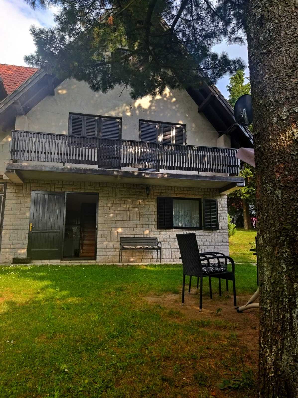A two-story house exterior features a stone facade and black shutters. A balcony overlooks a grassy area, with a black chair and small table positioned below. Sunlight filters through the branches of a nearby tree, creating a serene outdoor environment.