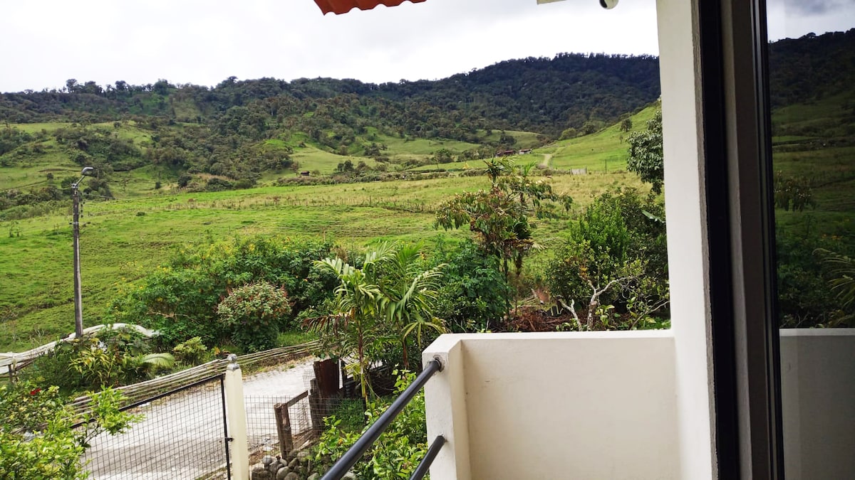 A view from the balcony showcases lush green hills and a tranquil landscape. Various plants and trees can be seen in the foreground, while a pathway leads into the distance, inviting exploration of the natural surroundings.