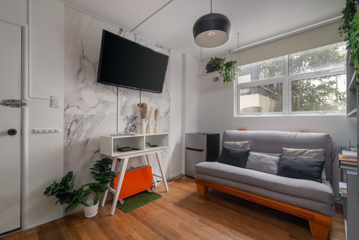 A bright living area features a gray sofa with decorative cushions, complemented by a white TV stand against a marbled wall. Natural light enters through large windows adorned with light-colored shades. Indoor plants add a touch of greenery to the space.