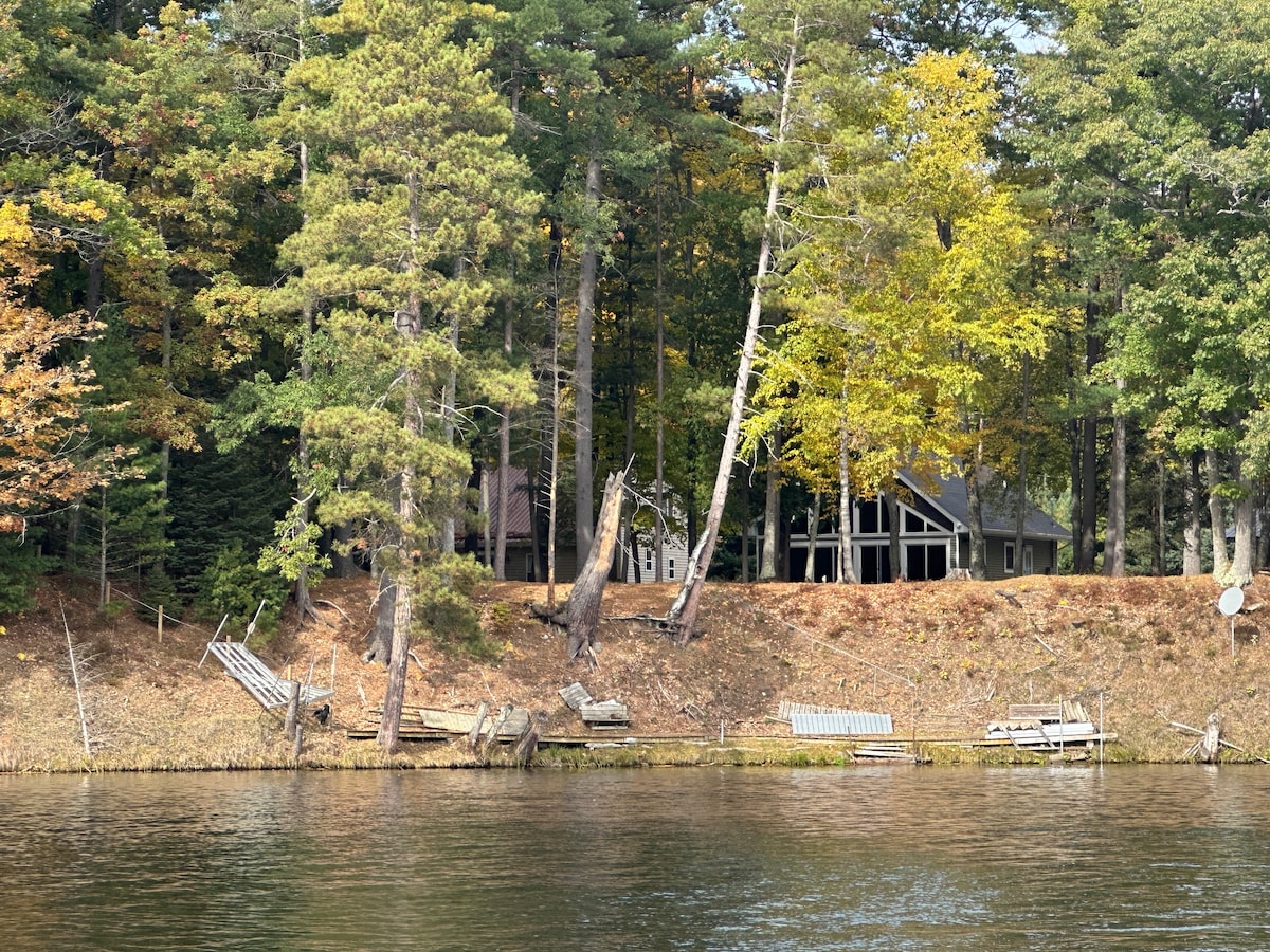 A serene view of a riverbank features a house nestled among tall trees. Several wooden docks are visible along the shoreline, showcasing a blend of natural beauty and outdoor access. The autumn foliage adds a touch of color to the tranquil setting.
