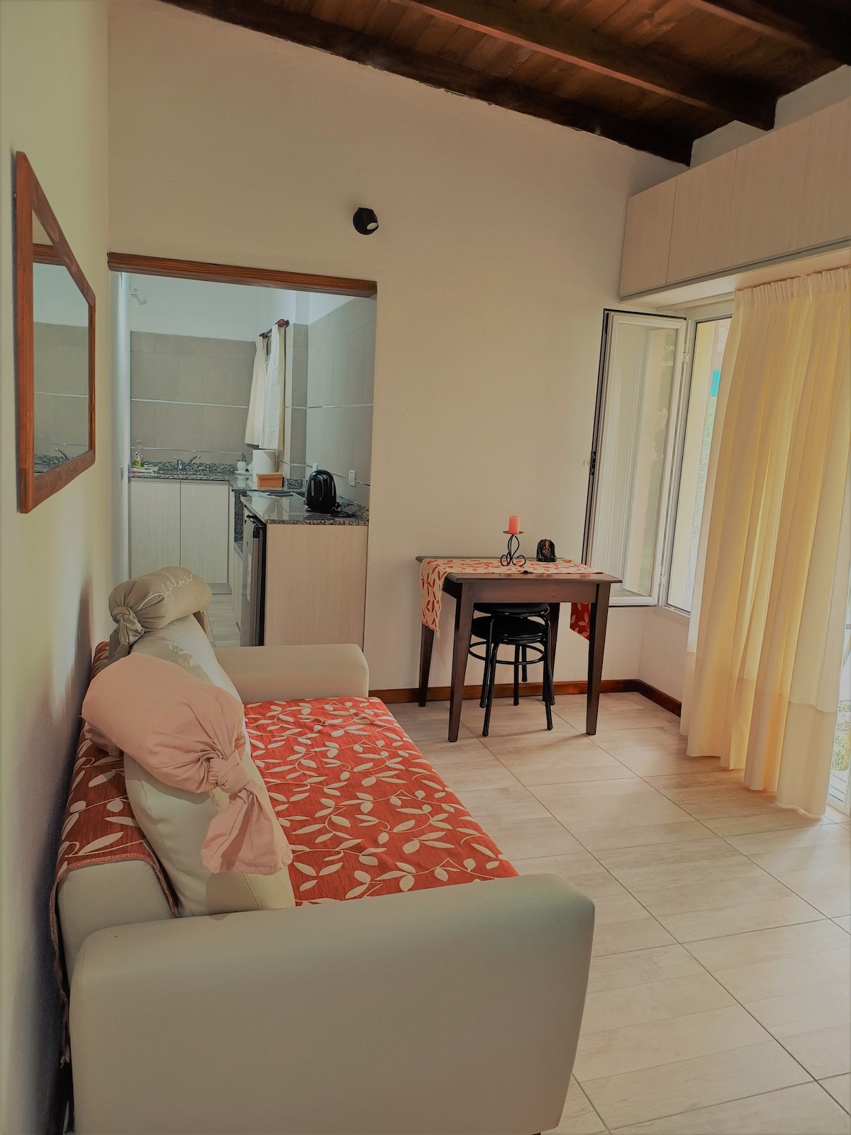 A cozy seating area features a white sofa adorned with a coral-patterned throw pillow. A small table and chair are positioned nearby, with a gentle view offered through the large window that is dressed in light curtains.