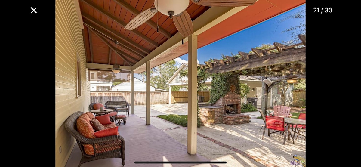 An outdoor space is featured with a covered porch supported by tall, wooden columns. A rustic brick outdoor fireplace is visible, surrounded by shaded seating. The pergola above adds charm, while the well-maintained yard provides a serene atmosphere.