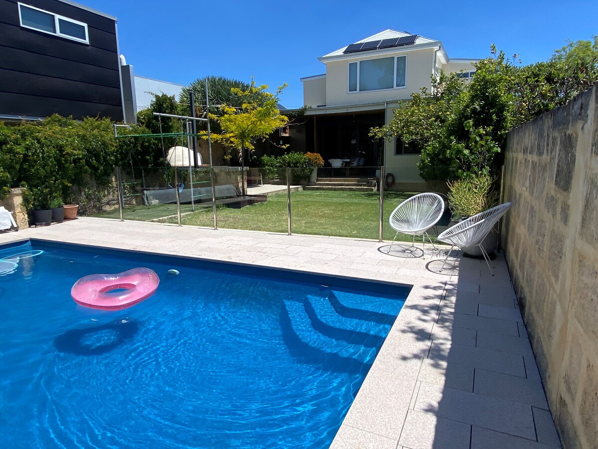A clear blue swimming pool is featured prominently in the foreground, with a pink inflatable ring floating on the water. Beyond the pool, a well-maintained lawn and patio can be seen, with a small tree and modern architecture framing the backdrop.
