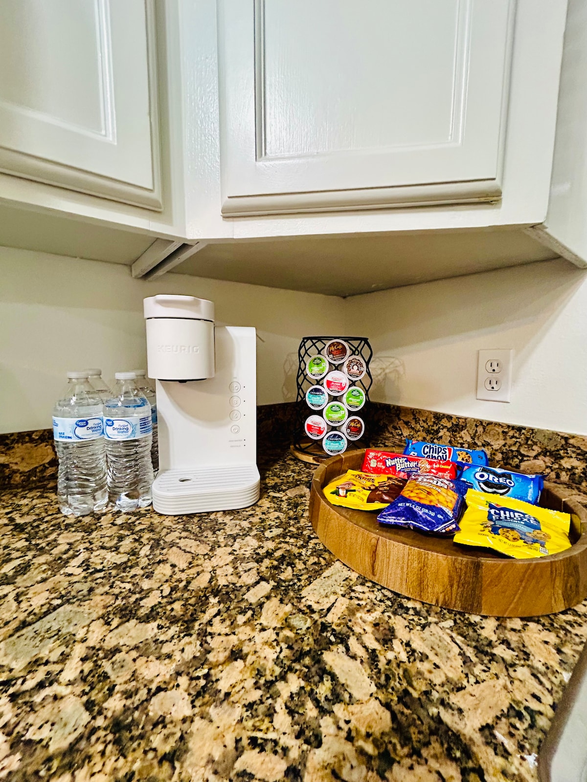 A kitchen corner displays a coffee maker alongside three bottles of water. A circular wooden tray holds an assortment of snacks, while a carousel holder features various coffee pods, providing options for beverages. The countertop showcases a classic granite pattern.