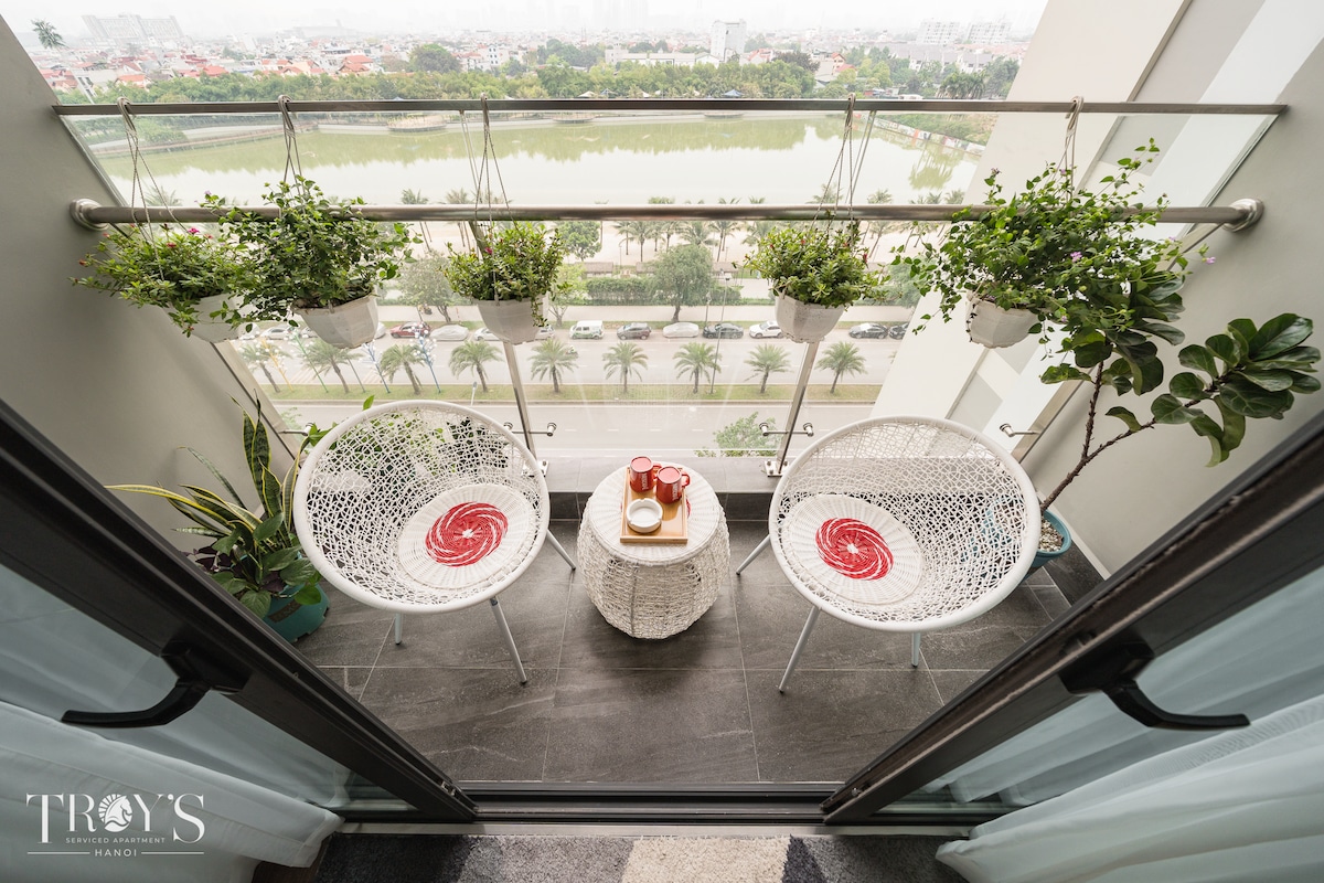 A balcony is equipped with two white, circular chairs and a small table. Decorative plants hang from the railing, while a panoramic view of the lake and greenery is visible in the background. Light drapes frame the doorway leading to the interior.