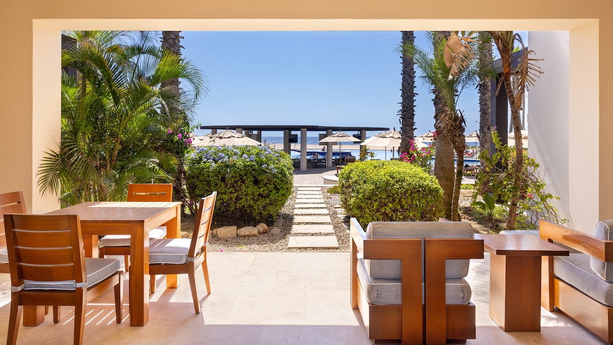A spacious outdoor patio features a wooden dining set and comfortable seating, framed by lush greenery and palm trees. The view opens to a beautifully landscaped area leading to a pool area with lounge chairs and umbrellas, allowing for a seamless blend of indoor and outdoor relaxation.