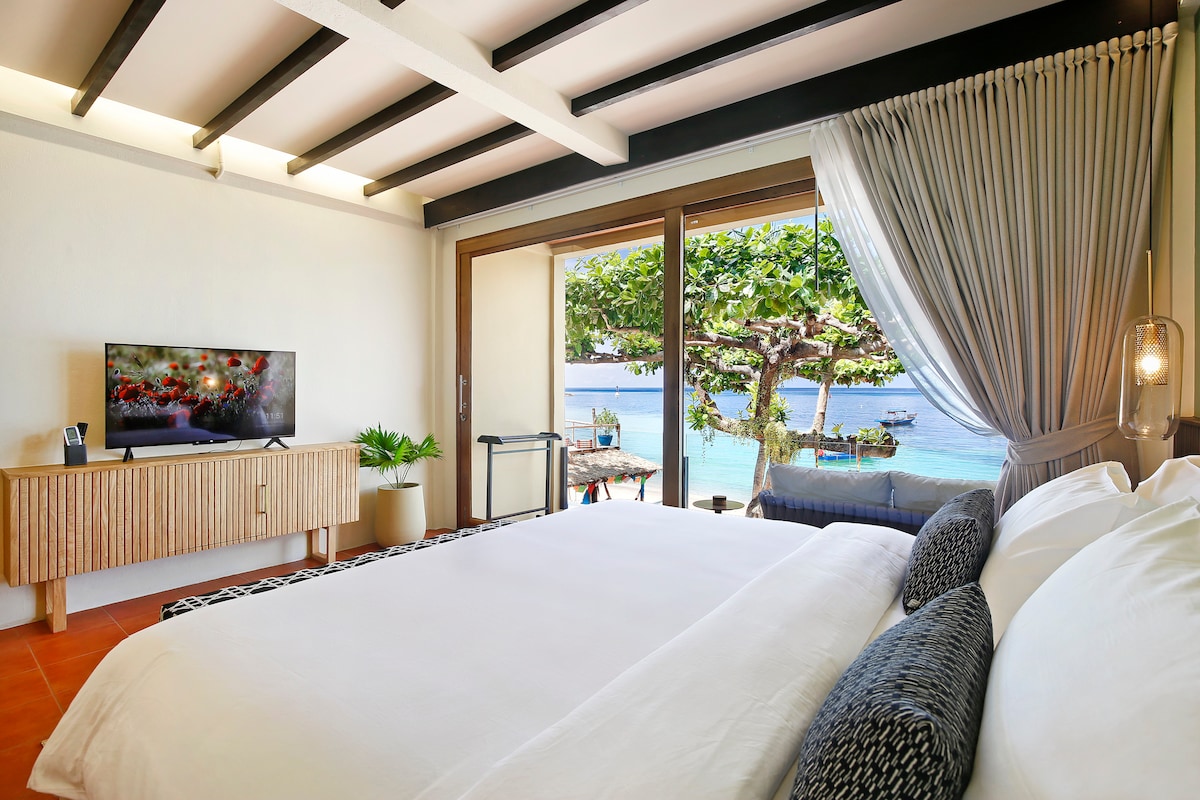 A bright studio room features a spacious bed with soft pillows and a view of the ocean through large sliding glass doors. A smart TV and a tasteful wooden console are present. Lush greenery and lounge seating are visible on the balcony.
