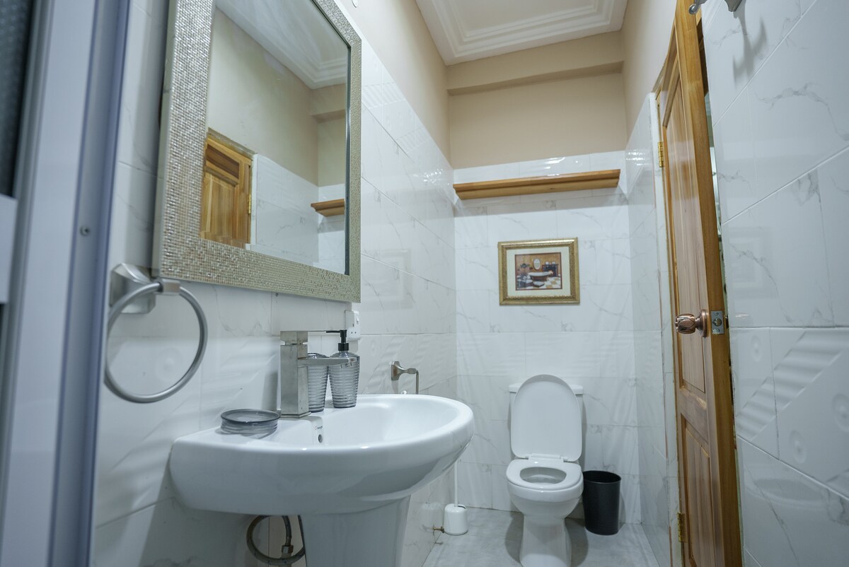 A well-maintained bathroom is presented, featuring glossy white tiles and softly illuminated by natural light. A modern sink with a large mirror is positioned above it, while a toilet sits next to a small wastebasket. A decorative framed artwork adds a touch of character to the space.