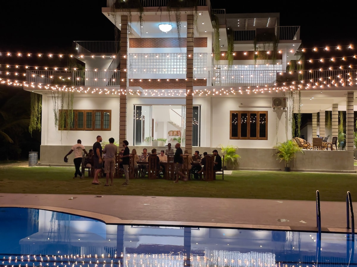 A spacious outdoor area is illuminated by string lights over a long dining table filled with guests. The modern building features large windows and a balcony, all reflected in the pool below. Lush greenery is positioned around the grounds, enhancing the vibrant atmosphere.