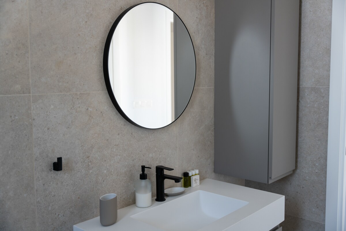 A modern bathroom features a sleek, round mirror above a stylish vanity. The countertop holds a selection of soap dispensers and a small cup. A gray cabinet is mounted on the wall, providing additional storage space. The walls are finished in a light stone texture.
