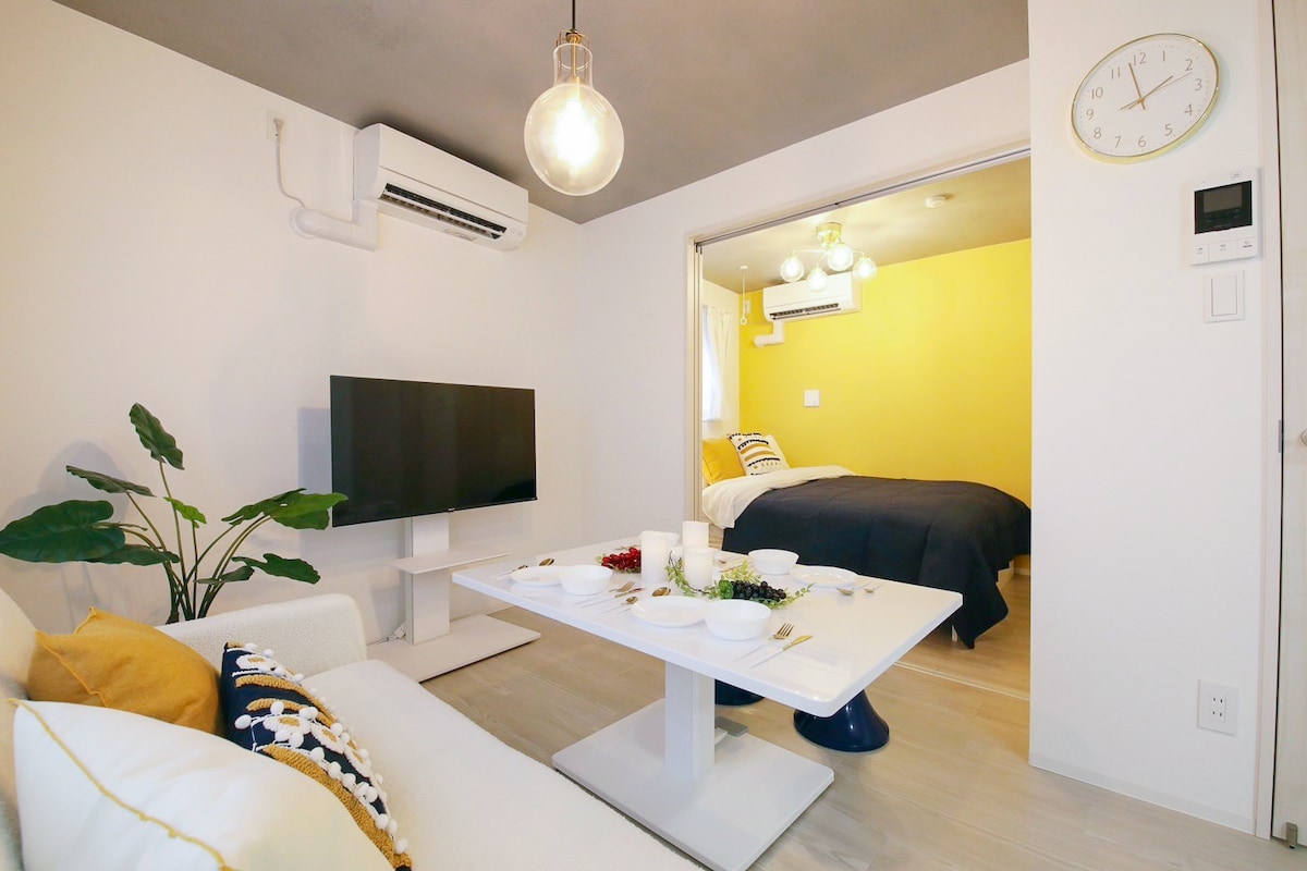 A modern living area features a white sofa adorned with decorative cushions. A sleek, round dining table is set with a variety of fruits, and a flat-screen television is mounted on the wall. The space is connected to a vibrant yellow bedroom, enhancing the overall brightness.