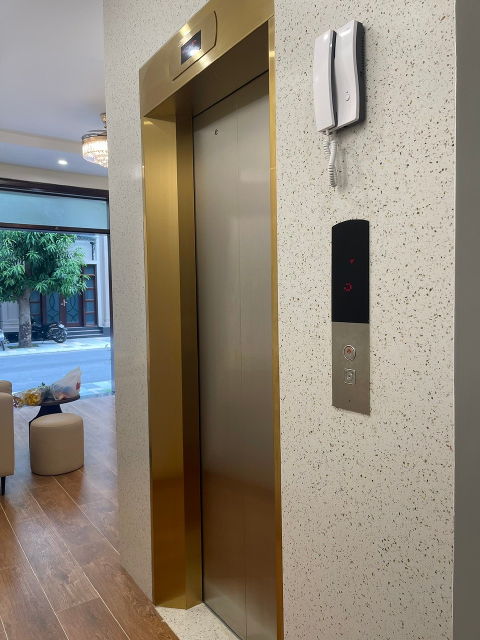 An elevator is positioned against a textured wall, featuring a polished gold door. A control panel, equipped with buttons and a intercom system, is mounted beside the elevator. Soft lighting creates an inviting atmosphere in the surrounding area, with views of greenery visible through glass doors.