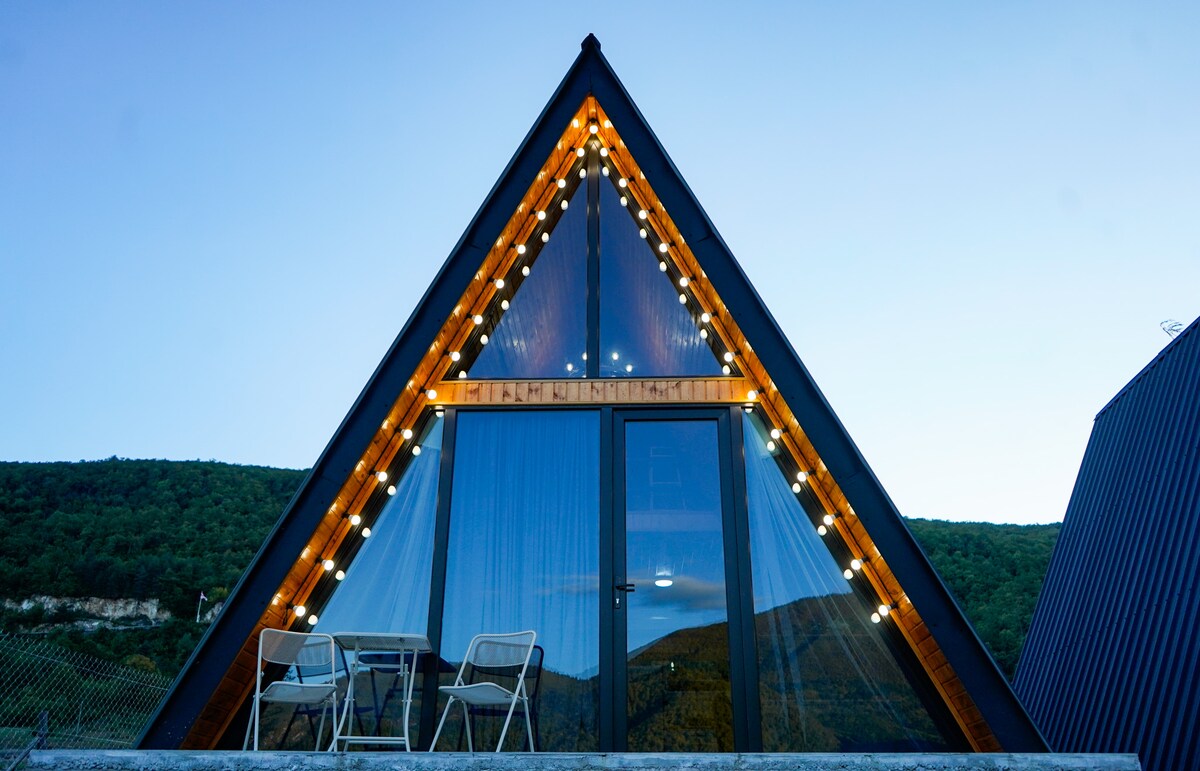 The exterior of a triangular cottage features large glass doors framed by wooden accents. Soft lighting is strung along the roof edge, creating a welcoming appearance. A small balcony is visible with two chairs, offering a view of the surrounding landscape.