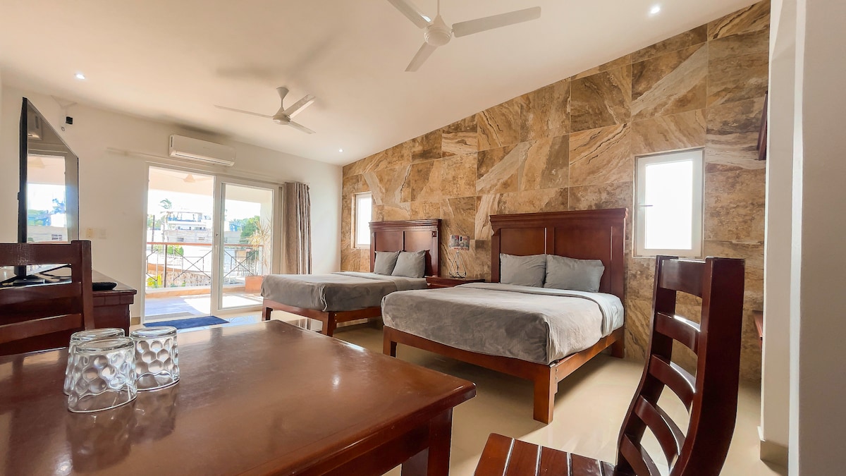The spacious room features two queen beds with soft gray bedding, a wooden table and chairs, and a large wall mounted television. Natural light enters through two windows and glass doors that lead to a terrace, with a warm stone wall adding texture.