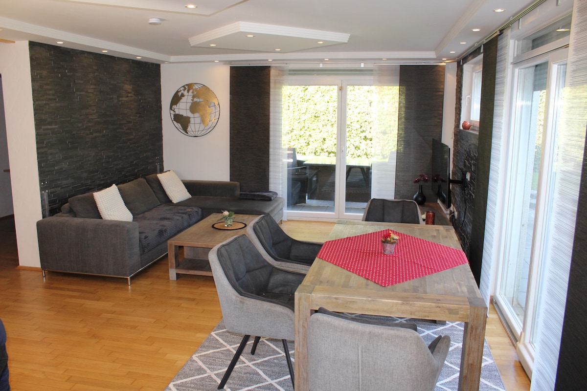 An open living area features a modern gray sofa and two accent chairs surrounding a wooden dining table. Large glass doors offer access to an outdoor terrace, while stone accent walls add a textured backdrop. Decorative elements include a globe wall art piece and a vibrant red table runner.
