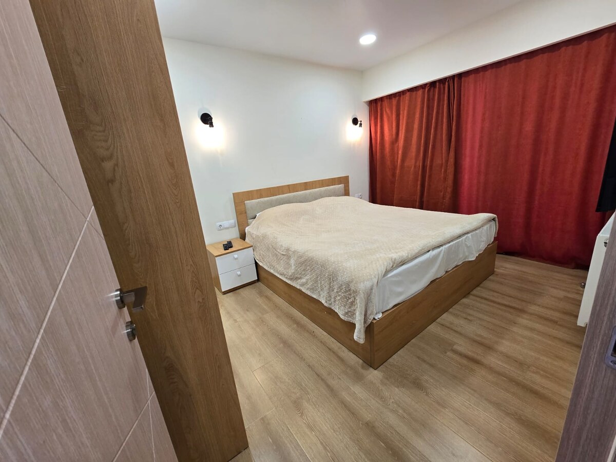 A calming bedroom features a spacious bed with a light-colored blanket and wooden headboard. The room is enhanced by soft lighting and wall sconces, complemented by a rich red curtain. A nightstand is positioned beside the bed, while natural wood flooring adds warmth.