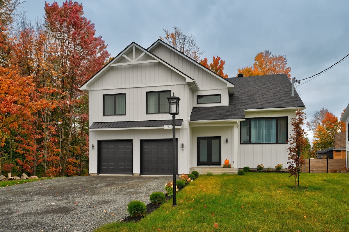 A modern two-story house is set against a backdrop of vibrant autumn foliage. The front yard features neatly trimmed bushes, with a pathway leading to a welcoming entrance, highlighted by decorative pumpkins. The house is characterized by large windows and a two-car garage.