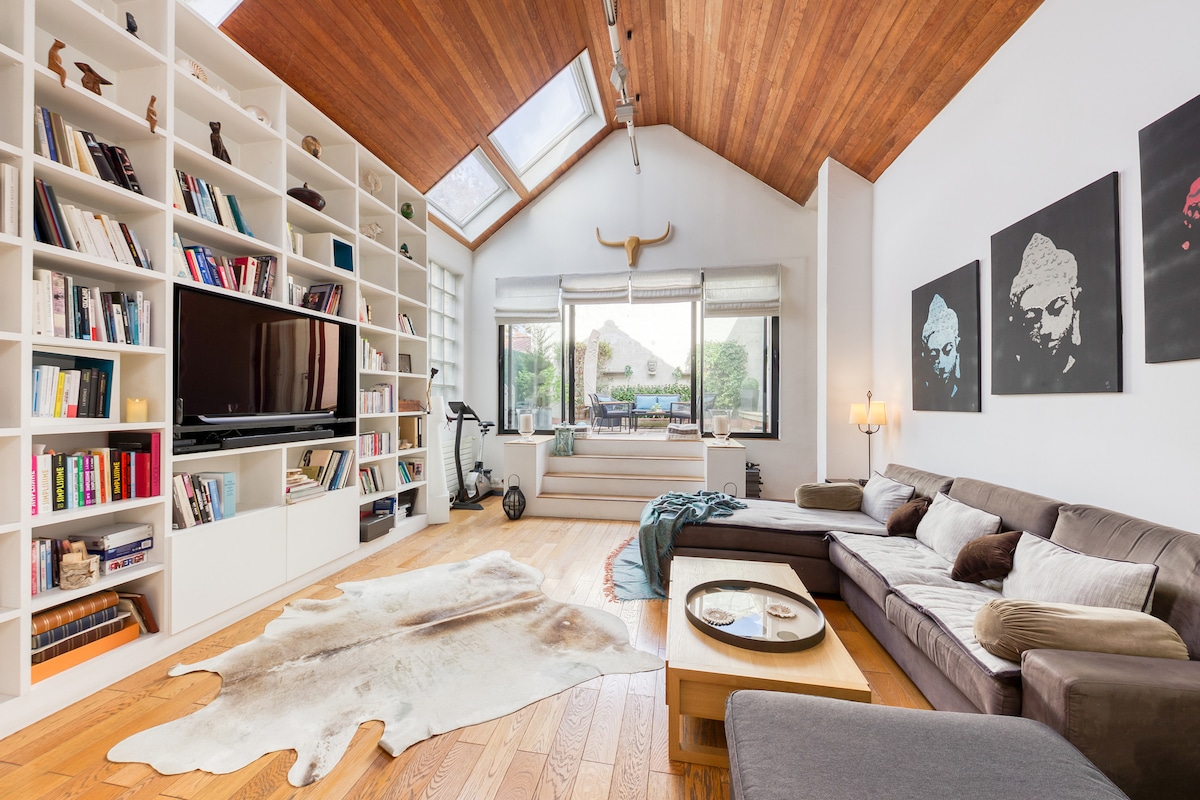 A spacious living area features a large sectional sofa and a coffee table, complemented by a natural cowhide rug. Floor-to-ceiling bookshelves line one wall, while large windows allow ample natural light, revealing views of greenery outside. A wall-mounted TV is positioned for relaxation.