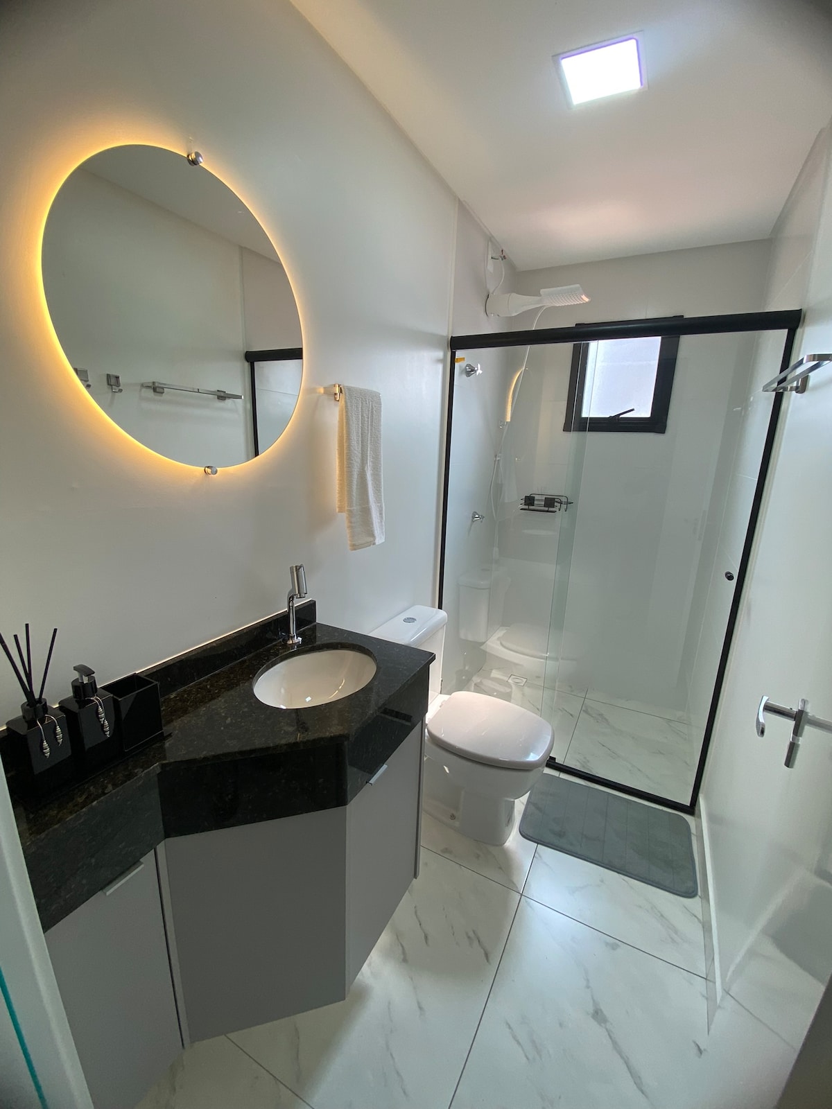 A modern bathroom features a stylish circular mirror with integrated lighting. A sleek black countertop is paired with a white sink, while a glass shower enclosure showcases a rainfall showerhead. The space is brightened by a small window and clean, white walls.