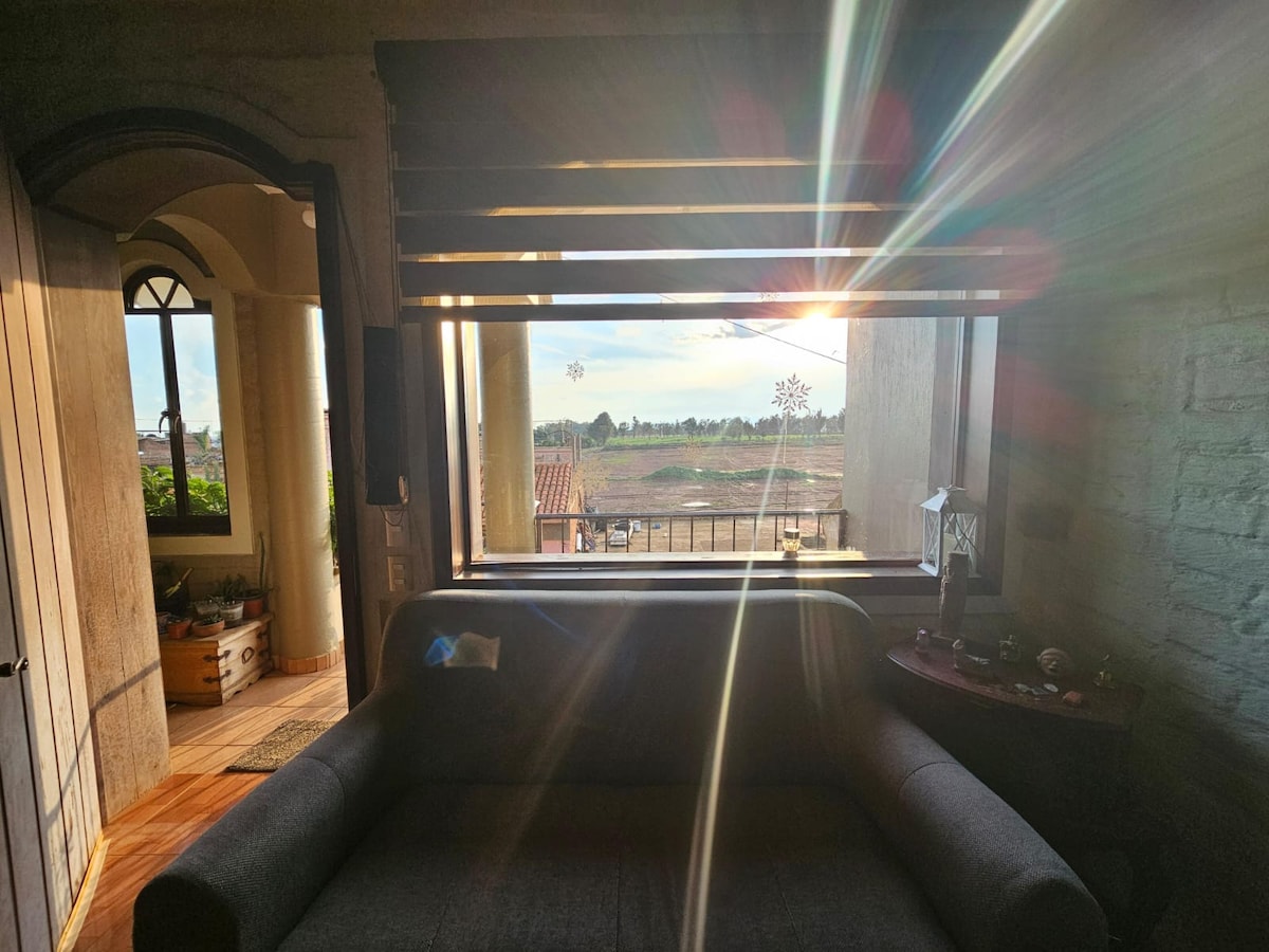 A cozy interior space is framed by a large window, allowing natural light to stream in. A dark sofa is positioned in front of the window, which overlooks a serene outdoor landscape illuminated by the sun. Subtle decor elements are arranged on a nearby table.