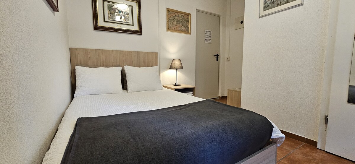 A cozy sleeping area features a spacious bed with white linens and a dark blanket. A bedside lamp provides soft lighting, complementing the neutral walls adorned with framed artwork. A door leads to the corridor, enhancing accessibility within the room.