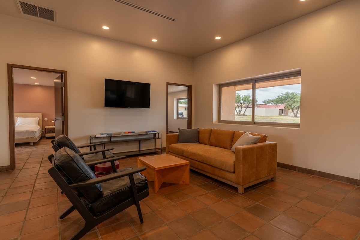 A spacious living area is showcased, featuring a cozy sofa and two armchairs. A wooden coffee table sits in the center, while a wall-mounted television is positioned for viewing. Natural light enters through the large window, illuminating the tile floor.