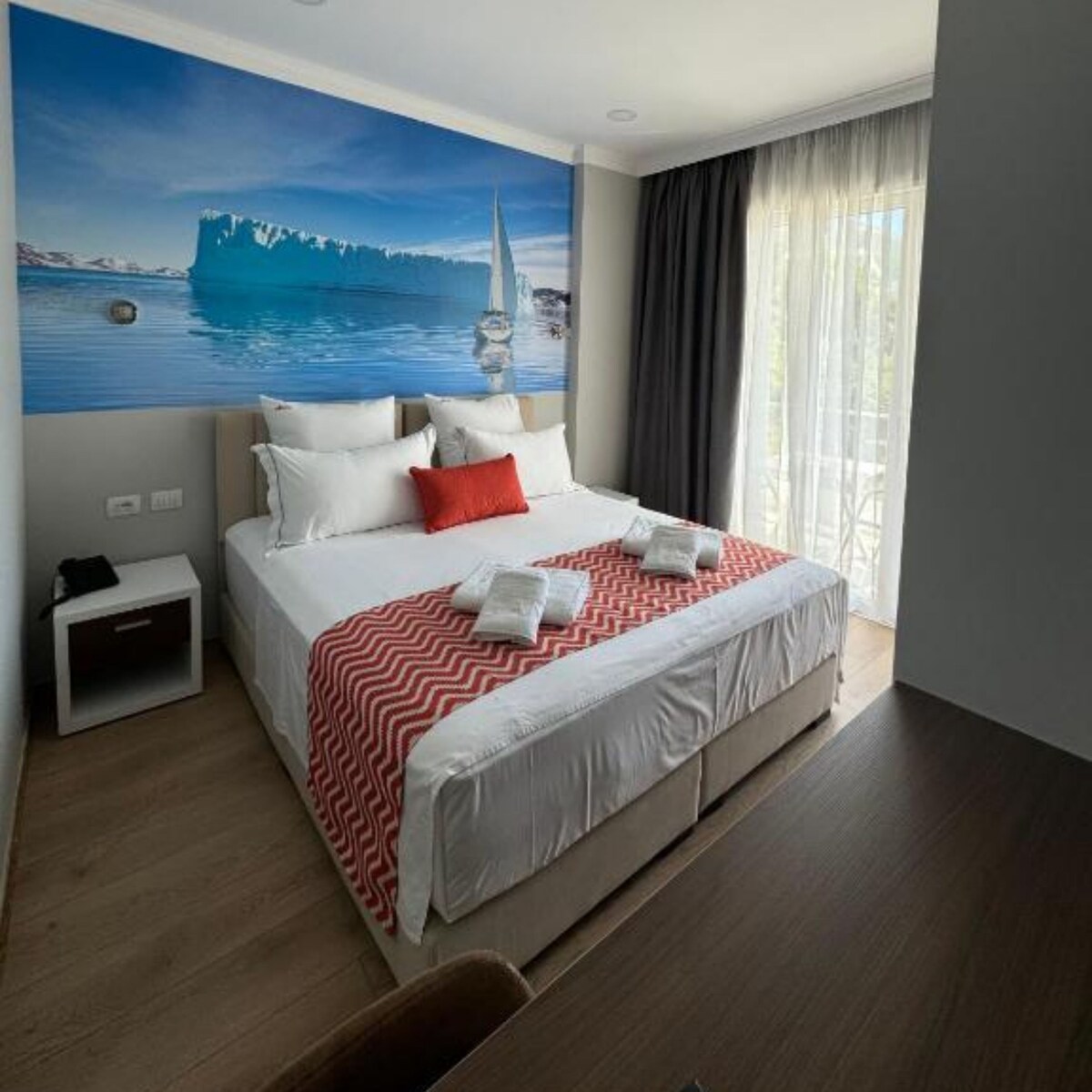 A bright bedroom features a spacious bed with crisp white linens and decorative pillows, including a vibrant red accent pillow. A large mural of an icy landscape adorns one wall, while natural light flows in through a window dressed with sheer curtains.