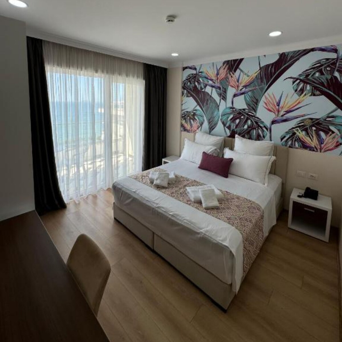 A bright bedroom features a spacious bed adorned with vibrant linens and cushions, complemented by tropical wall art. Large windows with sheer drapes allow natural light to fill the room, highlighting the wooden flooring and a small bedside table.