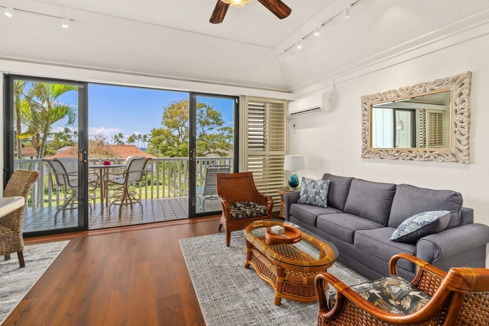 A bright living area features a comfortable grey sofa and two woven chairs, arranged around a round coffee table. Sliding glass doors open to a private balcony, revealing a view of trees and outdoor seating. Light wood flooring complements the space, illuminated by natural light.