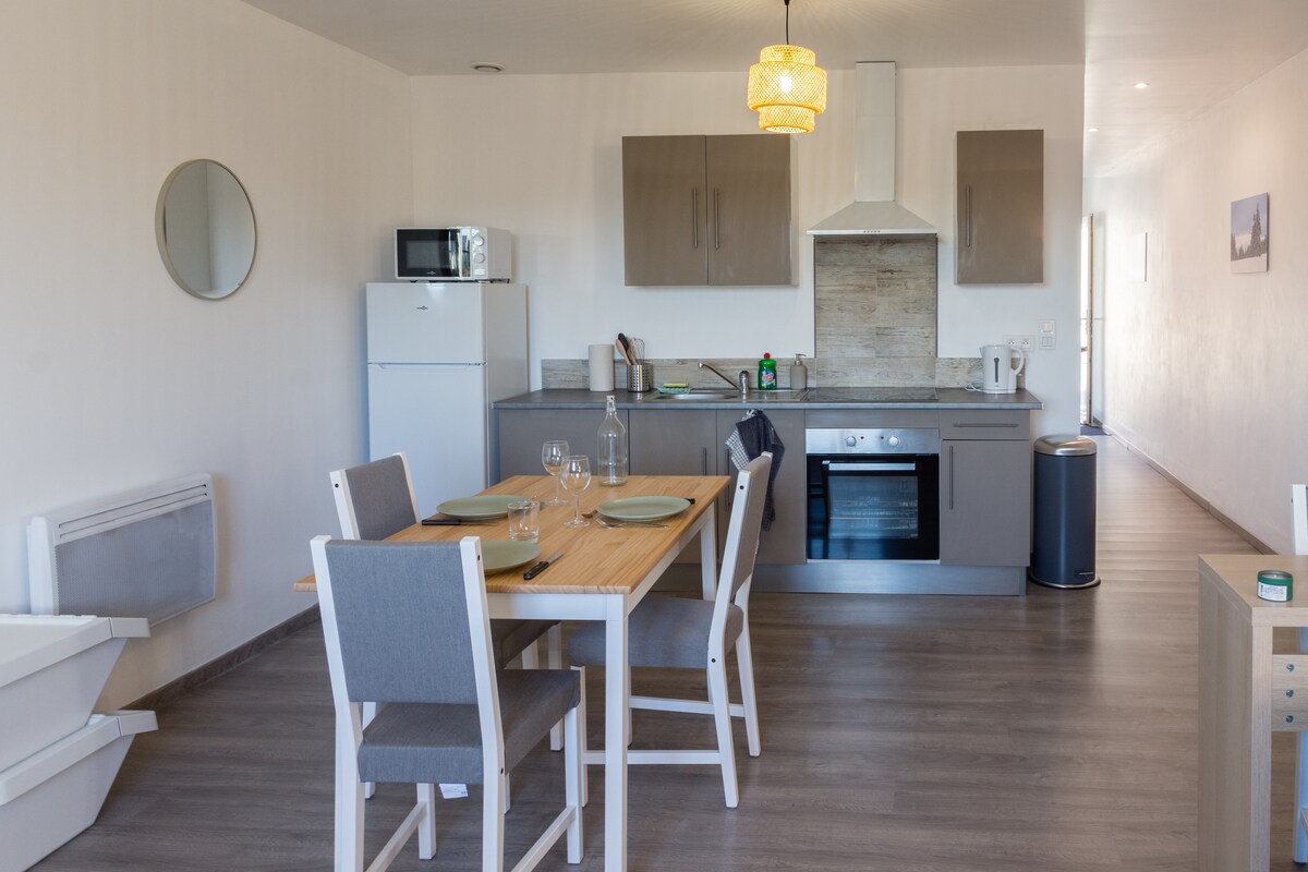 A functional kitchen and dining area is featured, equipped with modern appliances including a refrigerator and oven. A round dining table is set with plates and glasses, while light-colored cabinets provide ample storage. A large mirror reflects the space, enhancing the room's brightness.