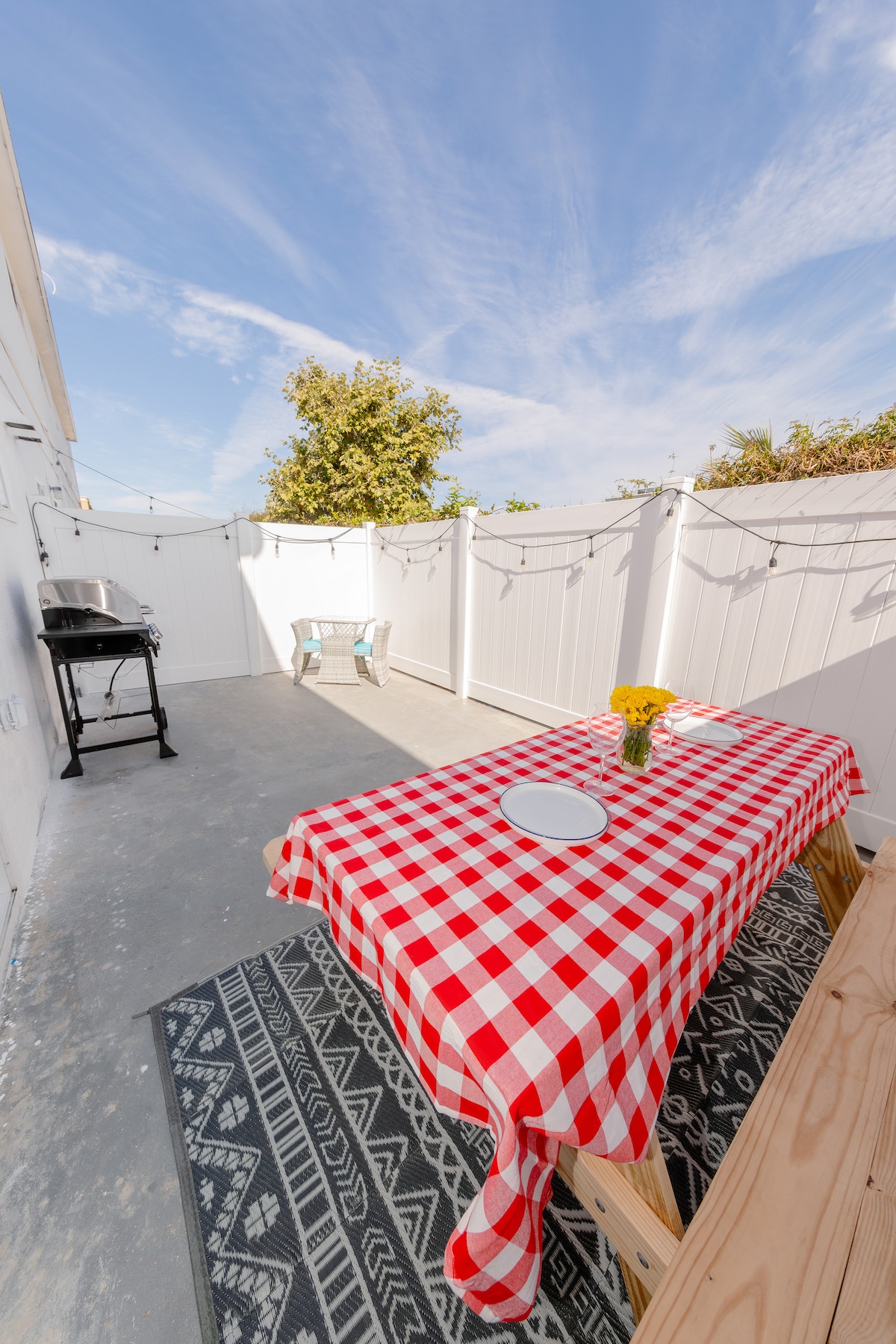 A private patio is showcased with a red and white checkered tablecloth covering a table, complemented by a vase of yellow flowers. A black grill is visible nearby, and string lights add a touch of charm under the bright blue sky.