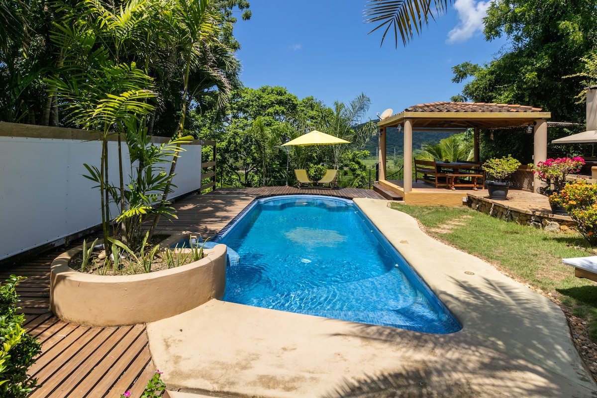 A private swimming pool is surrounded by landscaped greenery, featuring a sun deck with wooden flooring. A shaded terrace with seating is visible in the background, providing a relaxing space to enjoy the natural surroundings under clear blue skies.