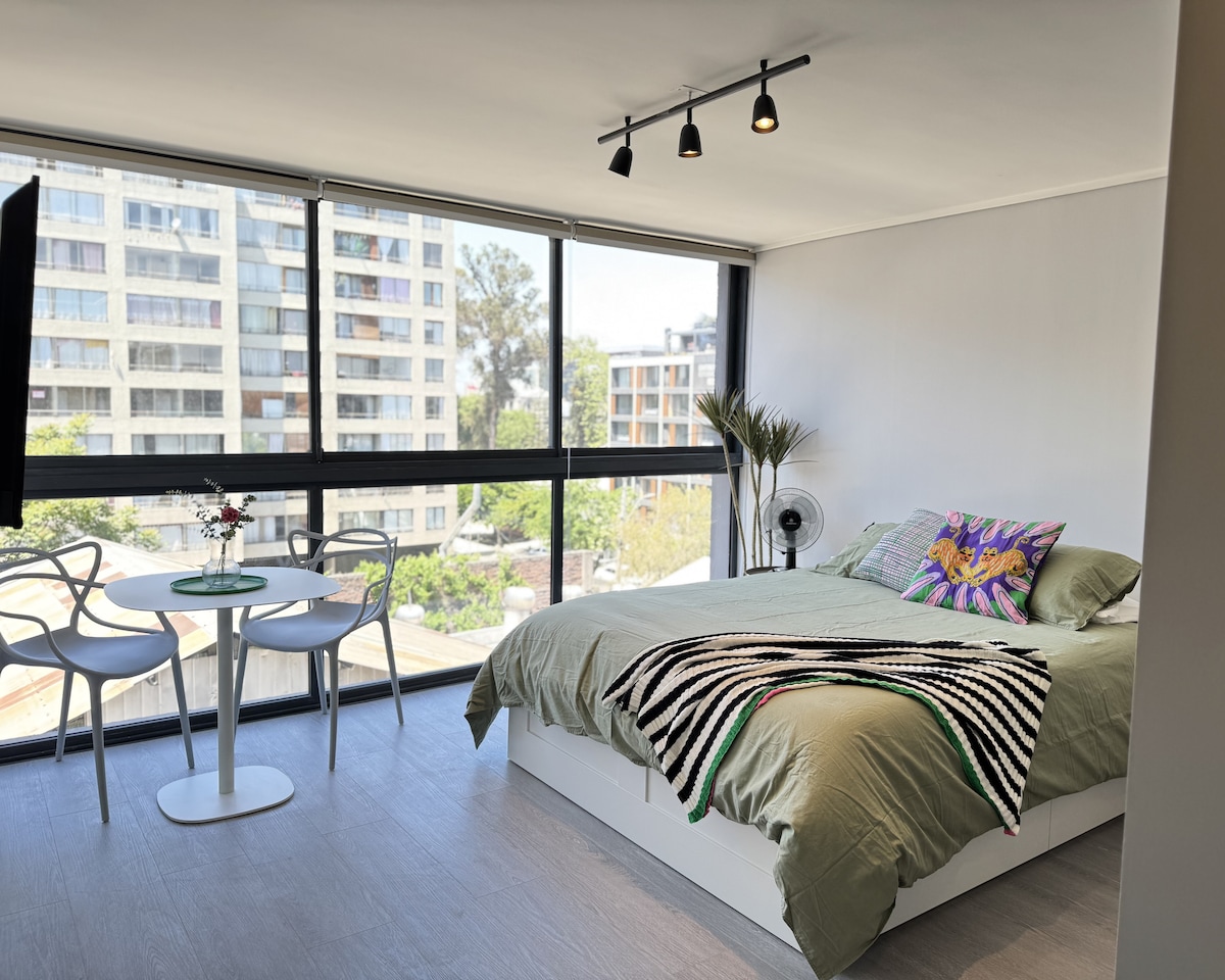 A bright studio space is filled with natural light from large windows, showcasing a cozy bed adorned with a striped blanket and decorative pillows. A small round dining table with two chairs is positioned nearby, offering a comfortable area for meals or relaxation.