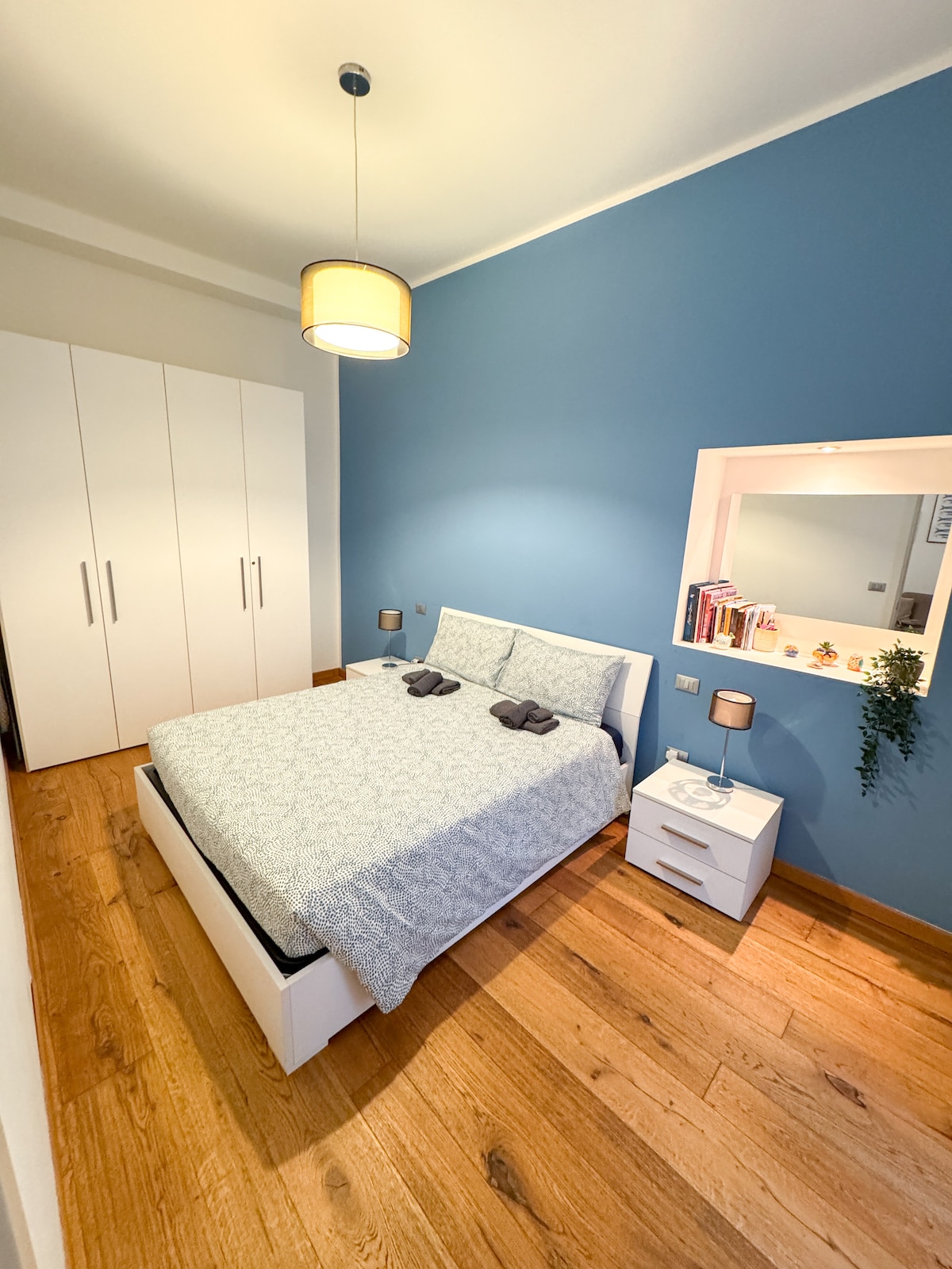 A serene bedroom showcases a spacious bed dressed in a light blue patterned blanket. A small bedside table with a lamp and a bookcase is visible, alongside a large wardrobe. Light hardwood flooring complements the calming blue accent wall.
